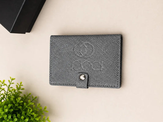A dark gray textured leather passport cover with an embossed '2030' design and a snap button closure, displayed on a light beige surface with a green plant and a black box in the background.