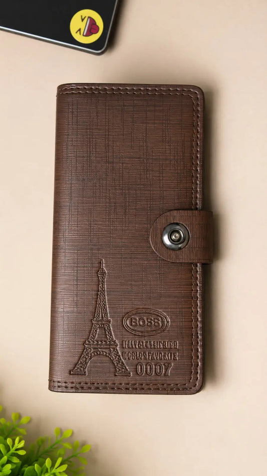A brown textured leather wallet with an embossed Eiffel Tower design and a snap-button closure, shown on a beige background with a small plant and a phone partially visible.